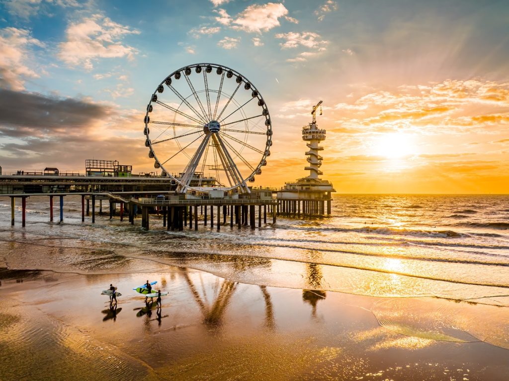 De Pier van Scheveningen voor zonsondergang. Drie surfers lopen op het strand met op de achtergrond een reuzenrad.