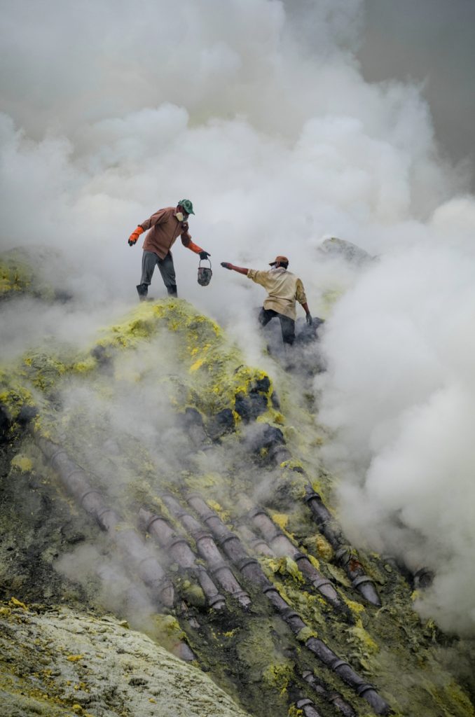 Twee mannen verzamelen zwavel tussen giftige dampwolken zonder voldoende bescherming. 
