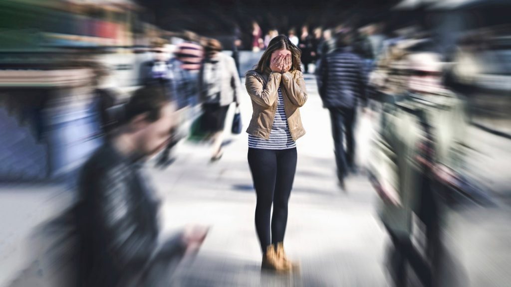 Jonge vrouw op drukke straat met handen voor ogen tijdens een paniekaanval. Ze heeft last van een angststoornis / anxiety.
