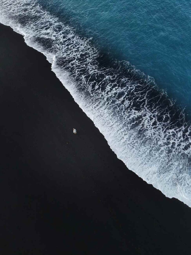 Topdown shot van persoon op zwart strand naast blauwe zee