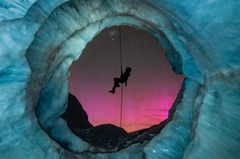 Foto vanuit een ijsgrot, waar iemand voor de opening abseilt en het zuiderlicht op de achtergrond