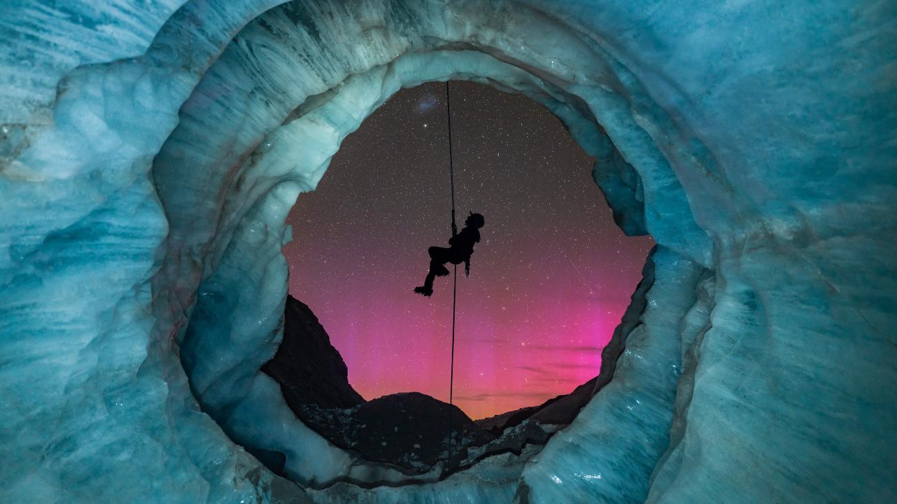 Foto vanuit een ijsgrot, waar iemand voor de opening abseilt en het zuiderlicht op de achtergrond