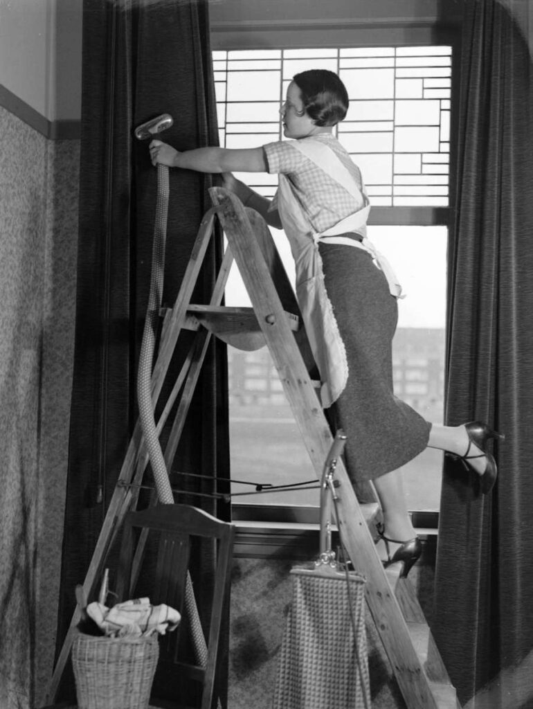 Vrouw in jaren zeventig maakt gordijnen schoon met stofzuiger terwijl ze op een trappetje staat